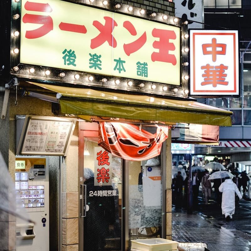 A lively Tokyo street scene with people passing by a ramen shop glowing on a rainy night.